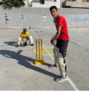 Cricket in Morocco: a batter in a red shirt and protective pads stands beside yellow stumps, with a wicketkeeper behind.